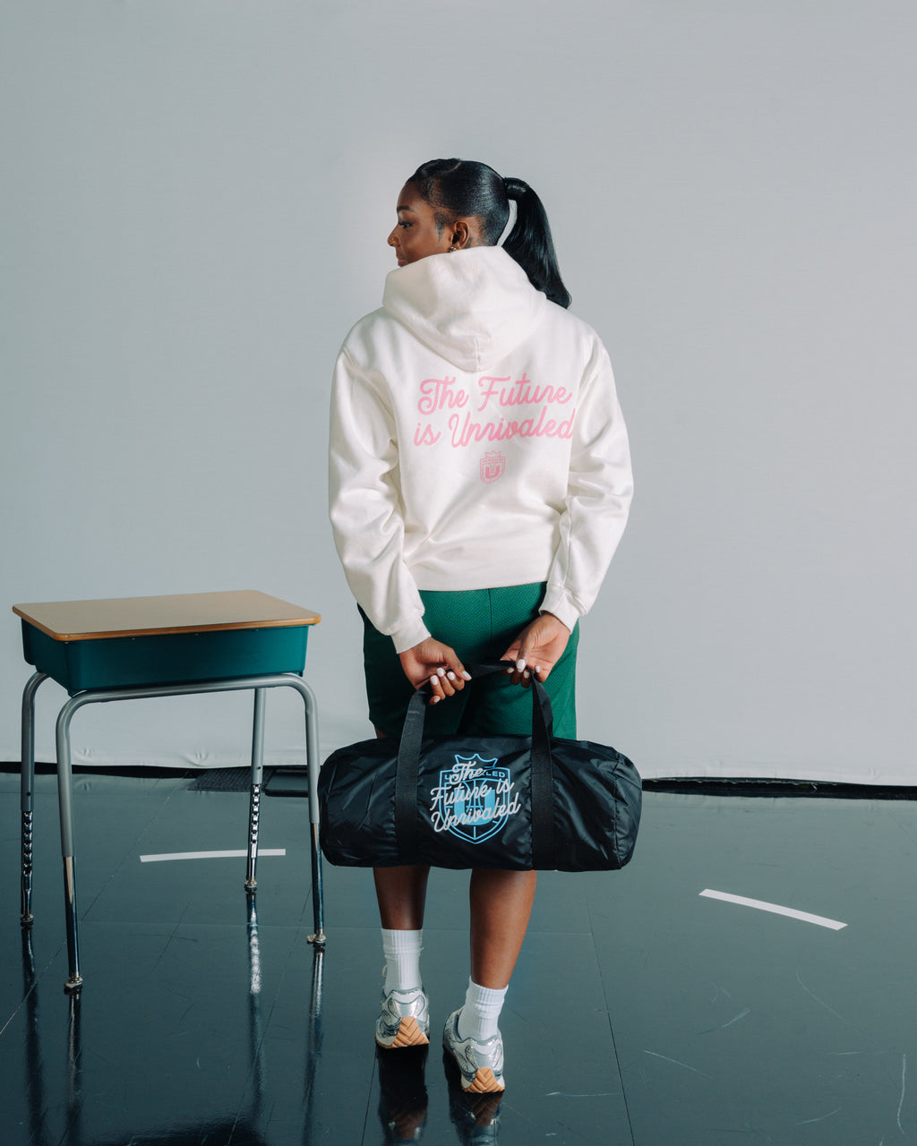 Ta’Niya Latson wearing a white hoodie with text, holding a black duffel bag in an indoor setting.