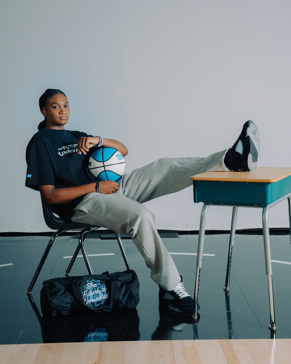 Madison Booker sitting on a chair with a basketball, wearing a black "The Future Is Unrivaled" shirt