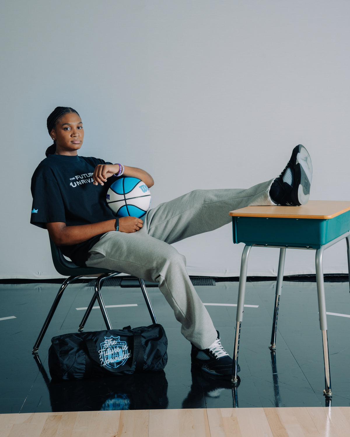 Madison Booker sitting on a chair with a basketball, wearing a black "The Future Is Unrivaled" shirt